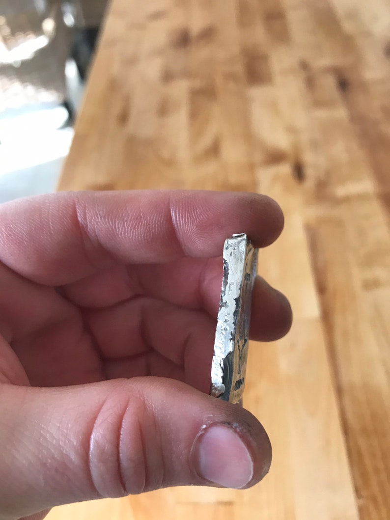 Puede incluir: Una barra de metal rectangular de color plateado con un borde rugoso y texturizado y una superficie estampada. La pieza de metal tiene un brillo met&aacute;lico y parece ser un lingote fundido con marcas visibles y lados irregulares.