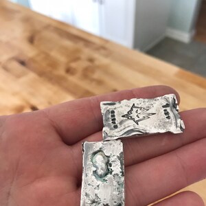 Puede incluir: Dos lingotes de plata rectangulares con una textura rugosa y fundida a mano. Uno presenta un logotipo de rayo estampado y el n&uacute;mero 1.89. El otro tiene una superficie irregular y picada con oxidaci&oacute;n visible y color plateado met&aacute;lico.