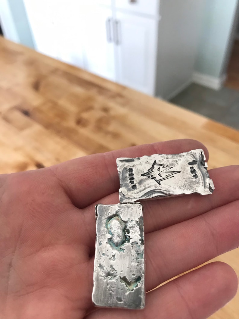 Puede incluir: Dos lingotes de plata rectangulares con una textura rugosa y fundida a mano. Uno presenta un logotipo de rayo estampado y el n&uacute;mero 1.89. El otro tiene una superficie irregular y picada con oxidaci&oacute;n visible y color plateado met&aacute;lico.