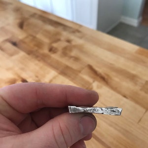 Puede incluir: Una peque&ntilde;a barra de metal rectangular de color plateado con una superficie rugosa y texturizada y bordes irregulares. El objeto tiene un brillo met&aacute;lico y una apariencia s&oacute;lida y densa, con un acabado bruto y sin pulir.