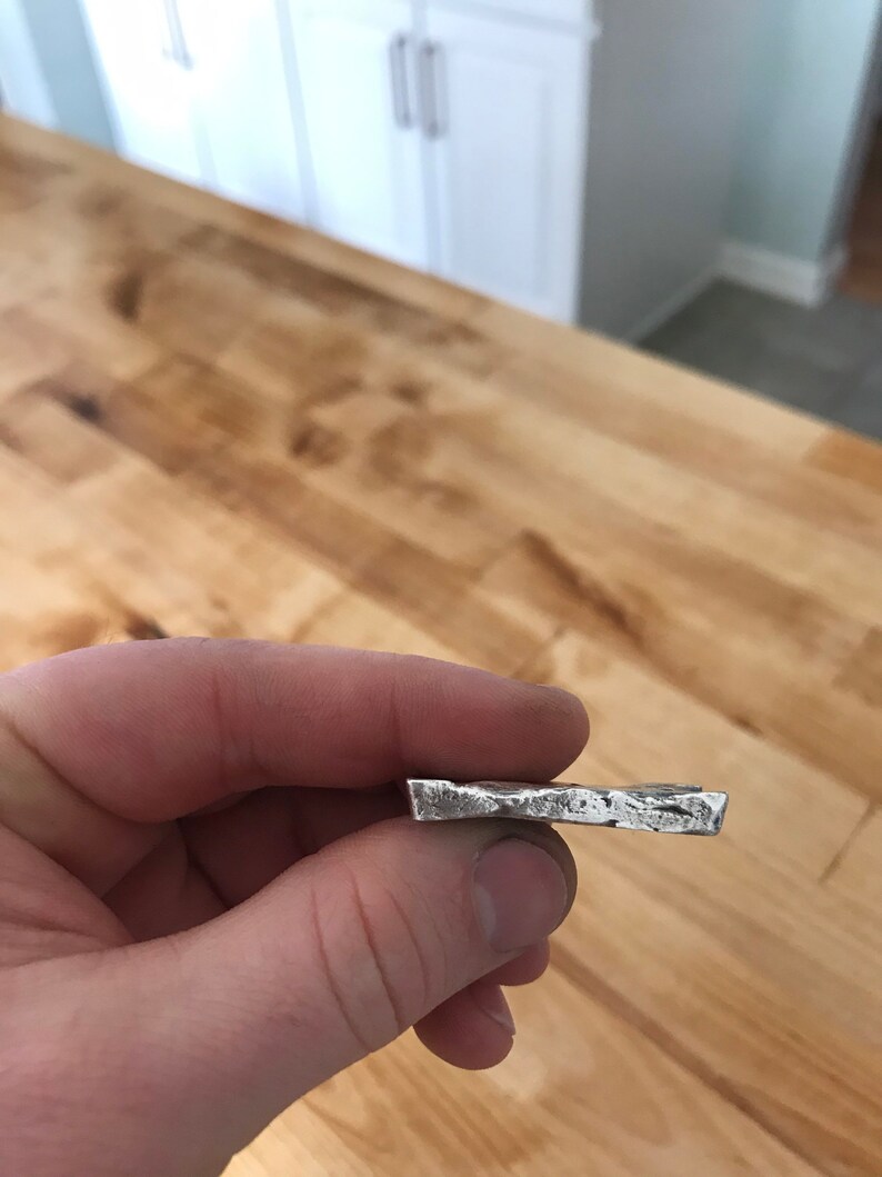 Puede incluir: Una peque&ntilde;a barra de metal rectangular de color plateado con una superficie rugosa y texturizada y bordes irregulares. El objeto tiene un brillo met&aacute;lico y una apariencia s&oacute;lida y densa, con un acabado bruto y sin pulir.