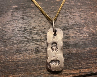 Puede incluir: Un colgante rectangular de color plateado con una superficie texturizada e irregular que cuelga de una cadena dorada. El colgante presenta un símbolo de calavera y tibias cruzadas estampado sobre el texto 999FS, indicando la pureza de la plata.