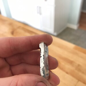 Puede incluir: Una barra de metal rectangular de color plateado con un borde texturizado e irregular y un acabado met&aacute;lico. El lado de la barra muestra texto estampado que incluye los n&uacute;meros 999 y otros caracteres parcialmente visibles.