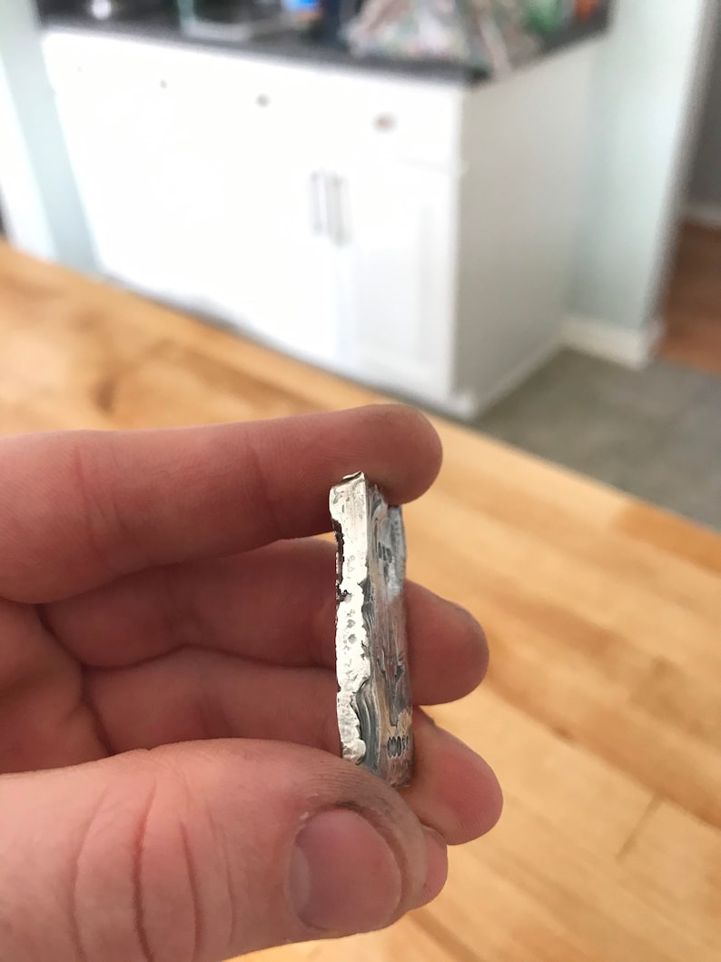 Puede incluir: Una barra de metal rectangular de color plateado con un borde texturizado e irregular y un acabado met&aacute;lico. El lado de la barra muestra texto estampado que incluye los n&uacute;meros 999 y otros caracteres parcialmente visibles.