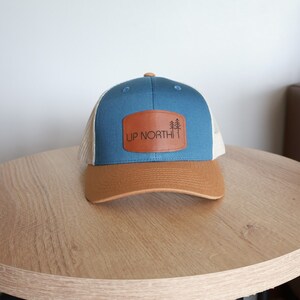 May include: A blue and tan trucker hat with a brown leather patch. The patch features the text "UP NORTH" and a graphic of three pine trees. The hat has a curved brim and a mesh back.