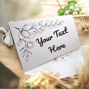 May include: A white rectangular folded greeting card featuring a black line art illustration of a floral branch on the left side. The text Your Text Here is printed in a black script font on the center of the card, which is partially tucked into an envelope.