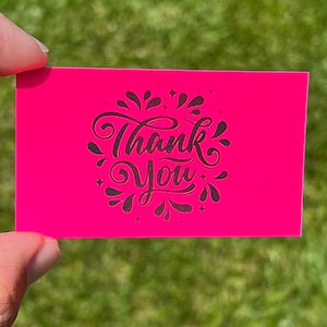 May include: A bright pink card with the words "Thank You" in black script lettering surrounded by a decorative design of black swirls and stars.