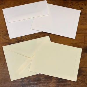 May include: Three blank envelopes, two white and one cream colored, with a subtle border around the edges. The envelopes are on a wooden surface.