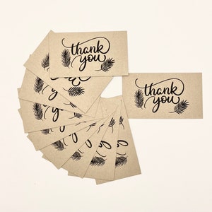 May include: A collection of kraft paper thank you cards. Each card features the words "thank you" in elegant black script, accompanied by fern-like illustrations. The cards are arranged in a fan shape, with several cards visible.
