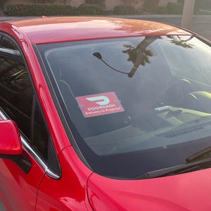 May include: A red car with a DoorDash delivery in progress sign on the windshield. The sign is white with a red D and the text "DoorDash Delivery in Progress".