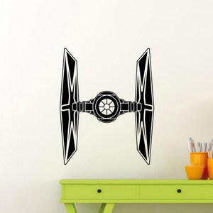 May include: Black and white wall decal of a TIE fighter, a fictional spacecraft from a science fiction franchise. The decal features a central circular cockpit with two angled wing panels extending outwards. The design is a silhouette.