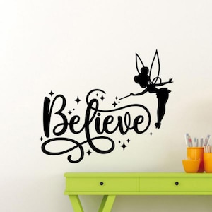May include: Black and white wall decal featuring a silhouette of Tinkerbell with her wand and the word "Believe" in a whimsical font. The decal is shown on a white wall with a light green dresser underneath.