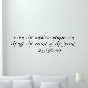 May include: Black script quote on a white wall: "Even the smallest person can change the course of the future. Lady Galadriel." The quote is in an elegant, flowing font, suitable for home decor.