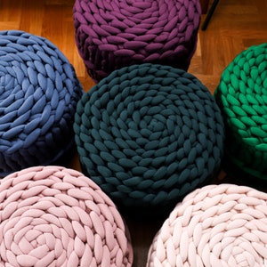 May include: Six round knitted poufs in different colors, including teal, purple, pink, and green. The poufs are made of thick yarn and have a textured surface.