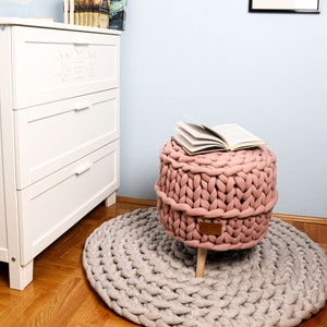 May include: A pink knitted pouf ottoman with wooden legs sits on a beige knitted rug. A book is placed on top of the ottoman.