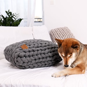 May include: A gray, chunky-knit pet bed with a leather tag sits on a white bed. A Shiba Inu dog with brown and white fur rests nearby. Additional knitted pillows are in the background. The bed is a cozy, stylish item for pets.