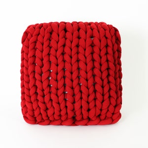 May include: A square, red, chunky-knit pouf. The knitted texture is prominent, with thick, braided strands creating a visually appealing pattern. The vibrant red color adds a bold accent to any room. This decorative piece can be used as a footstool or extra seating.