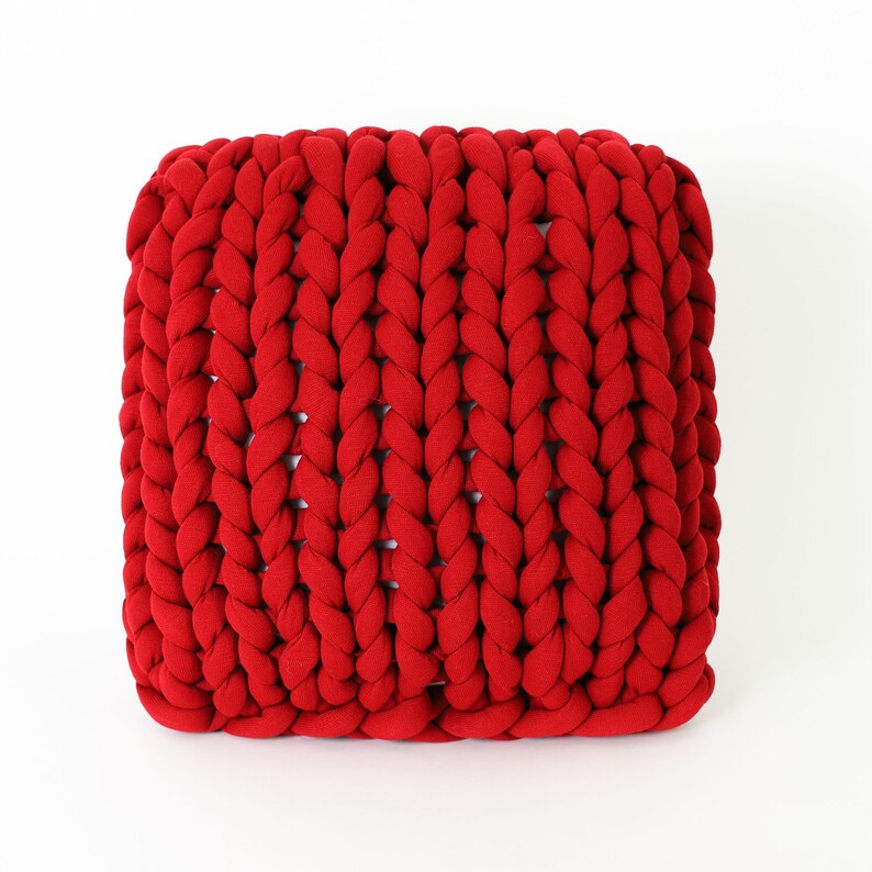 May include: A square, red, chunky-knit pouf. The knitted texture is prominent, with thick, braided strands creating a visually appealing pattern. The vibrant red color adds a bold accent to any room. This decorative piece can be used as a footstool or extra seating.