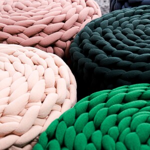 May include: Four round knitted poufs in different colors: pink, beige, dark green, and light green. The poufs are made of thick, chunky yarn and have a braided texture.