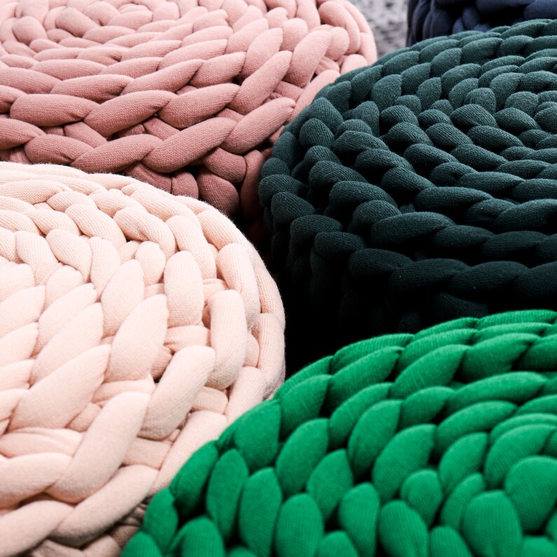 May include: Four round knitted poufs in different colors: pink, beige, dark green, and light green. The poufs are made of thick, chunky yarn and have a braided texture.