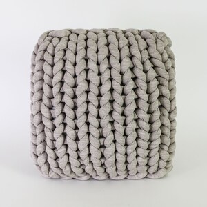 May include: A square, light gray pouf with a chunky, braided texture. The knitted design creates a visually appealing pattern, perfect for adding a touch of modern comfort to any living space. The neutral color allows it to blend seamlessly with various decor styles.