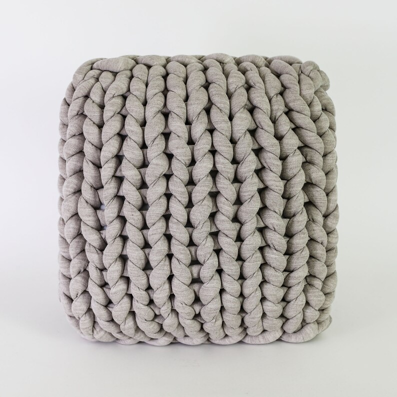 May include: A square, light gray pouf with a chunky, braided texture. The knitted design creates a visually appealing pattern, perfect for adding a touch of modern comfort to any living space. The neutral color allows it to blend seamlessly with various decor styles.