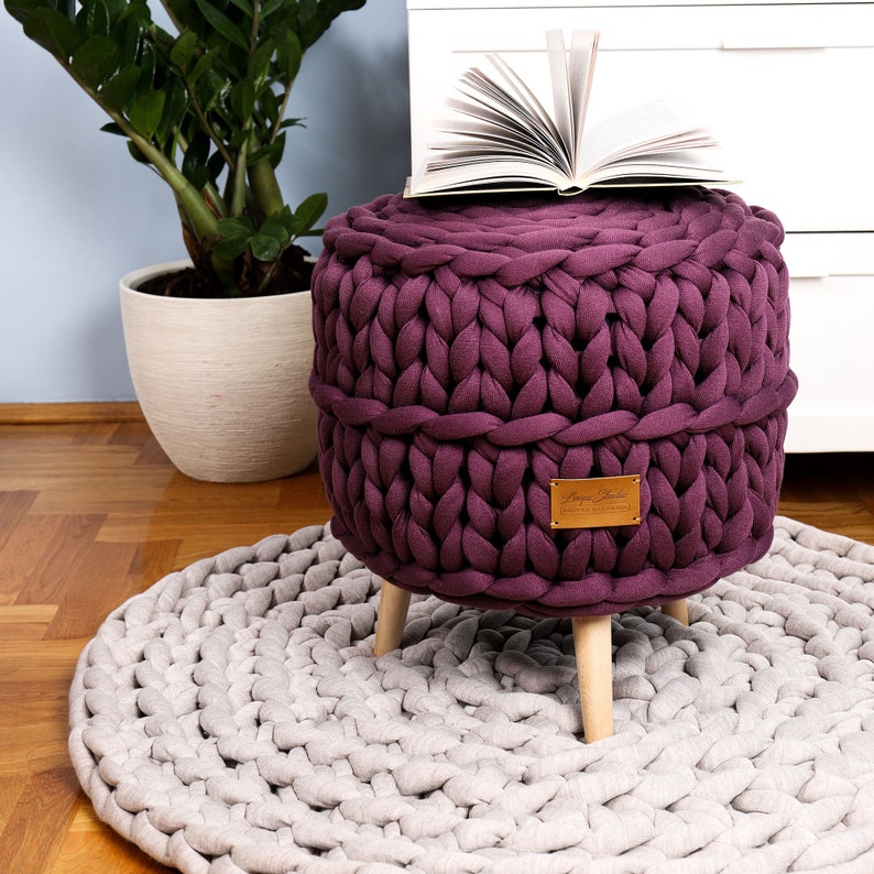 May include: A purple knitted ottoman with wooden legs sits on a light gray knitted rug. A book is placed on top of the ottoman.