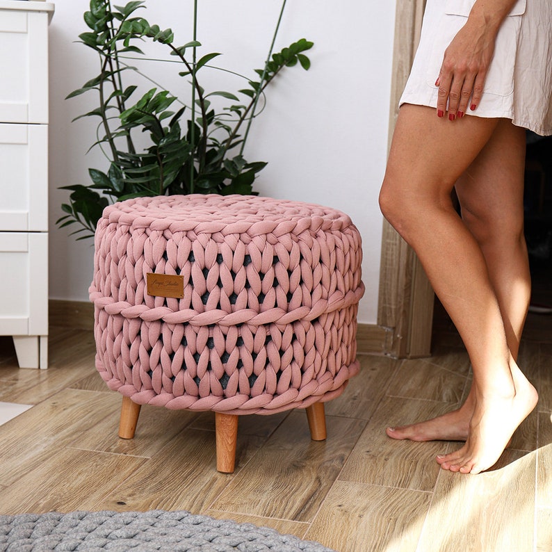 May include: A pink knitted ottoman with wooden legs. The ottoman is made of thick, chunky yarn and has a simple, modern design.
