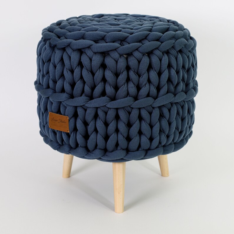May include: A navy blue knitted ottoman with a brown leather label that says "Loopy Studio". The ottoman has four wooden legs.