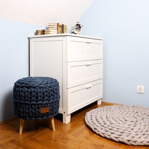 May include: A white dresser with three drawers and a blue knitted ottoman with wooden legs. A beige knitted rug is on the floor in front of the dresser.