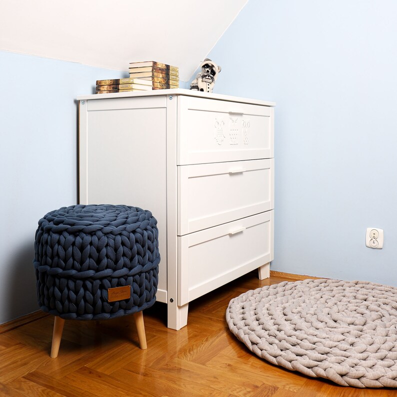 May include: A white dresser with three drawers and a blue knitted ottoman with wooden legs. A beige knitted rug is on the floor in front of the dresser.