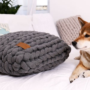 May include: A gray, chunky-knit dog bed with a brown leather tag. The bed is rectangular and sits on a white surface. A Shiba Inu dog is lying next to the bed.