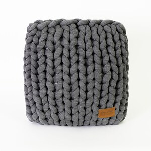 May include: A square, gray, chunky knit pouf. The knitted texture is visible, with thick, braided strands creating a textured surface. A small, brown leather tag is attached to the side. The pouf is a neutral color and sits on a white background.