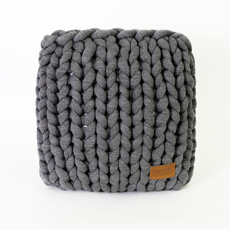 May include: A square, gray, chunky knit pouf. The knitted texture is visible, with thick, braided strands creating a textured surface. A small, brown leather tag is attached to the side. The pouf is a neutral color and sits on a white background.