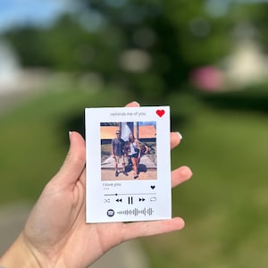 May include: A white card with a photo of two people, featuring the text "reminds me of you" and "I love you." The card has a Spotify-style music player design with a heart icon and play controls. The card is held in a person's hand.