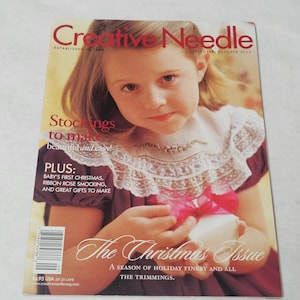 May include: A magazine cover featuring a young girl holding a wrapped gift. The magazine title is "Creative Needle" with the text "The Christmas Issue". The cover includes text about stockings, Christmas, and gifts.
