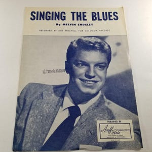 May include: Sheet music cover titled "Singing the Blues" by Melvin Endsley, featuring a portrait of a smiling man in a suit and tie. Published by Acuff-Rose Publications, with text indicating it was recorded by Guy Mitchell for Columbia Records.