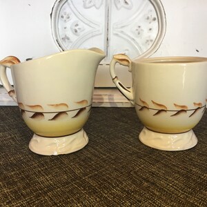 May include: A vintage cream-colored creamer and sugar set with brown leaf designs. The set has a yellow glaze on the bottom.