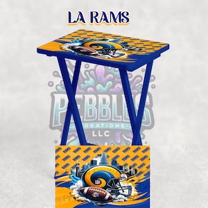 May include: A blue and yellow folding TV tray featuring the LA Rams logo and team colours. The tray has a blue frame and a tabletop with a repeating pattern of the team's logo and a football helmet graphic.