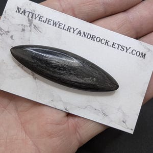 May include: A dark, elongated stone pendant with a polished finish, resting on a white card. The card features the text "NATIVEJEWELRYANDROCK.ETSY.COM" in a simple font. The pendant's shape is smooth and symmetrical, with a natural, textured appearance.