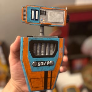 Op de afbeelding: Een blauw en oranje robot speelgoed met een scherm, knoppen en een compartiment met zilveren cilinders. De robot heeft een verweerde, verweerde look.