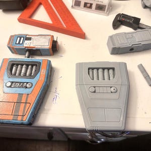 Op de afbeelding: Twee 3D-geprinte modellen van een futuristisch wapen. Eén is blauw, oranje en zwart geverfd, de andere is grijs. Beide modellen hebben een rij van zes cilinders en een bedieningspaneel met knoppen.