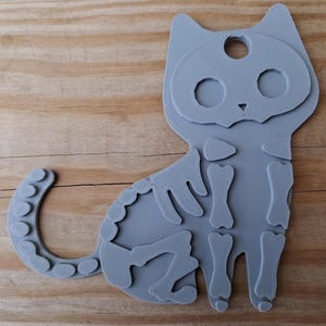 May include: A gray cat-shaped decoration with a skeleton design. The cat's body is outlined with a series of small circles, and the skeleton is visible within the cat's form. The cat has a hole at the top, suggesting it can be hung.