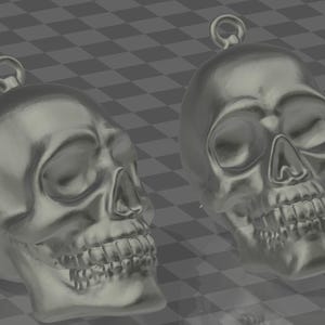May include: Two silver skull pendants, each with a loop for suspension. The skulls have detailed features, including eye sockets, a nose cavity, and teeth. The pendants are shown against a grey and white chequered background.