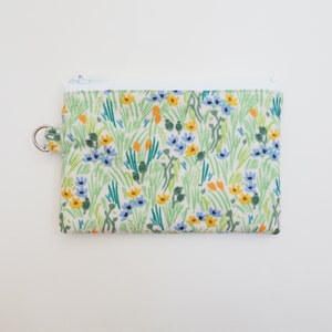 May include: A small zippered pouch with a white background and a floral print featuring blue, yellow, and green flowers. The pouch has a silver ring attached to the side.