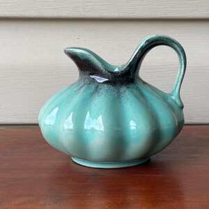 May include: A turquoise ceramic creamer with a dark brown rim and handle. The creamer has a bulbous body with vertical ridges and a curved handle. The creamer is sitting on a wooden surface.