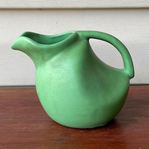 May include: A small, green ceramic pitcher with a rounded body and a curved handle. The pitcher has a spout for pouring and is likely used for serving liquids. The color is a muted, light green, and the pitcher appears to be made of a matte material.