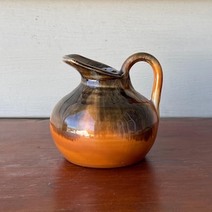 May include: A small, ceramic pitcher with a rounded body and a curved handle. The pitcher has a glossy finish with a gradient of colors, transitioning from a dark brown top to a vibrant orange base. The spout is slightly angled.