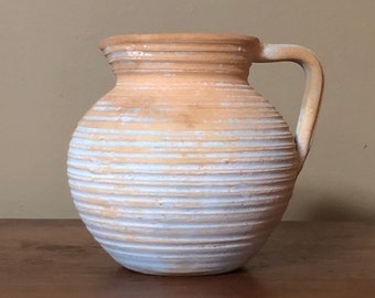 May include: A light brown ceramic pitcher with a white glaze and a ribbed design. The pitcher has a single handle.