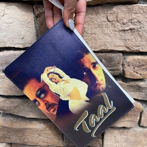 May include: Navy blue clutch with a white zipper and wrist strap. The clutch features a movie poster design with the word "Taal" in gold script. The design includes images of three people.